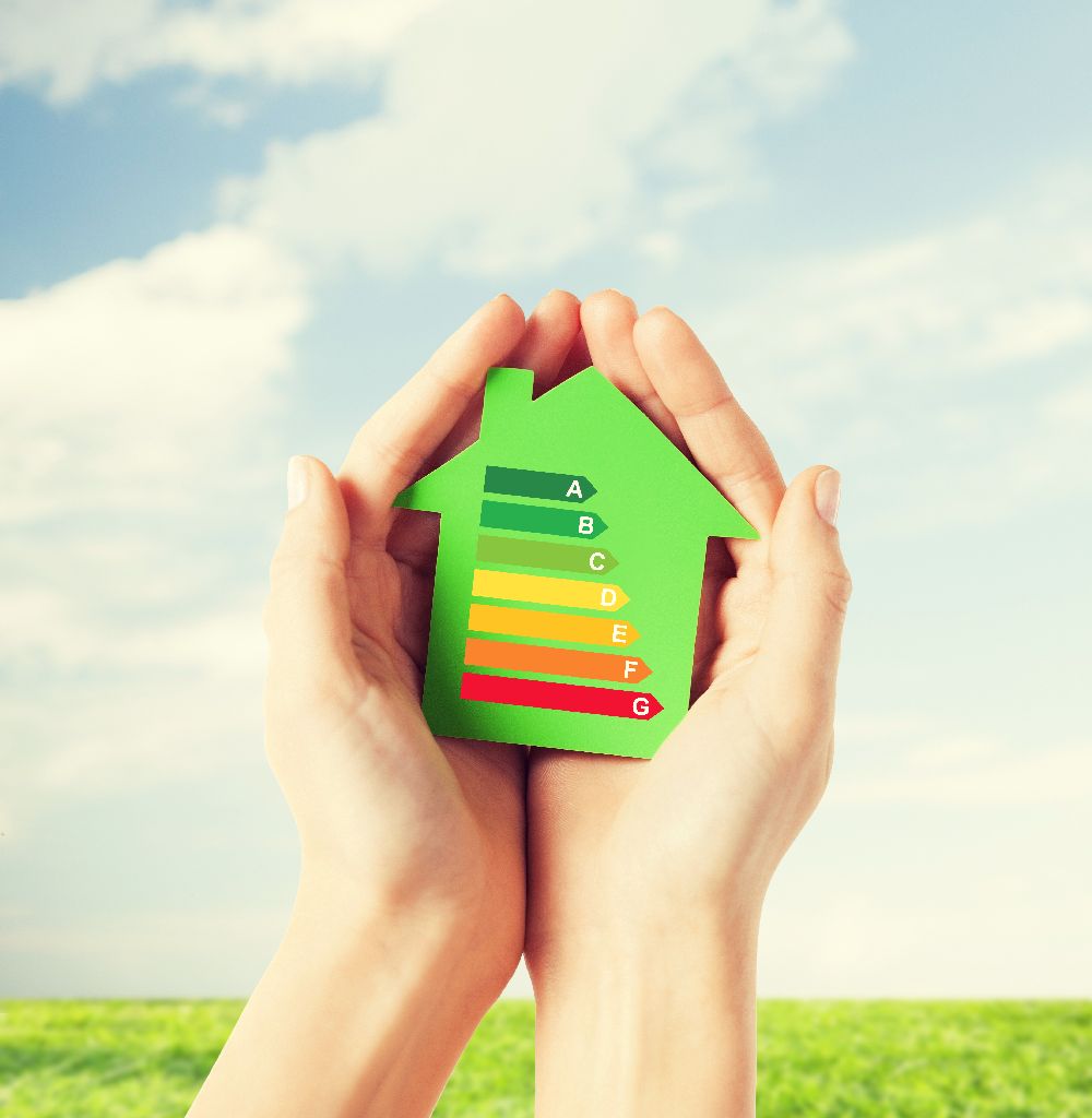 Manos unidas sosteniendo una madera verde con forma de casa, pintada con con un gráfico de categorías de ahorro energético, sobre fondo de hierba y cielo con nubes